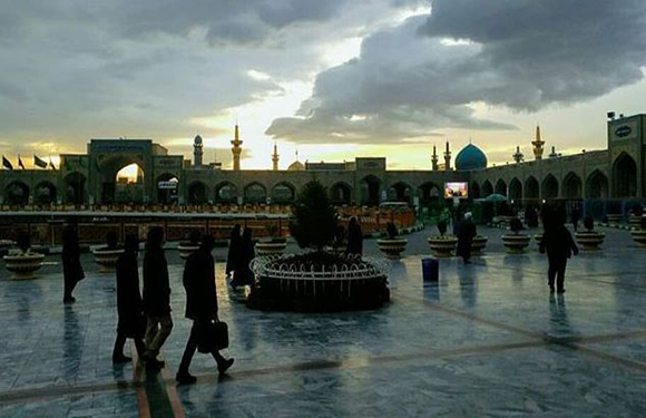 People traverse a wet courtyard with a fountain at dusk, mosque silhouettes and minarets visible against a cloudy sky.