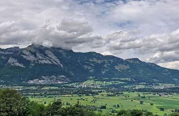 A scenic landscape with a mountain range in the background under a cloudy sky, and a lush green valley with scattered trees in the foreground.