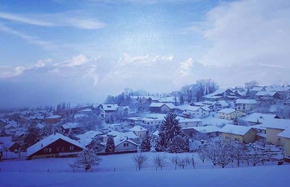 A snowy village with houses and trees covered in snow, under a blue-tinted sky with clouds partially obscuring the mountains in the background.