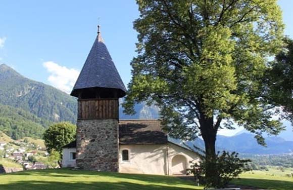 A small church with a conical roofed tower, adjacent to a large tree, set against a backdrop of mountains and a clear sky.