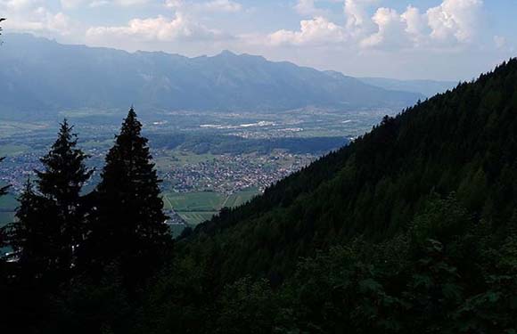 A landscape view overlooking a valley with a town, surrounded by mountains and forested areas, under a hazy sky.