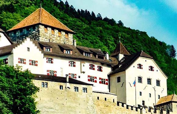 A traditional European castle with a stone tower and white walls with red window shutters, nestled on a hillside with lush green trees.