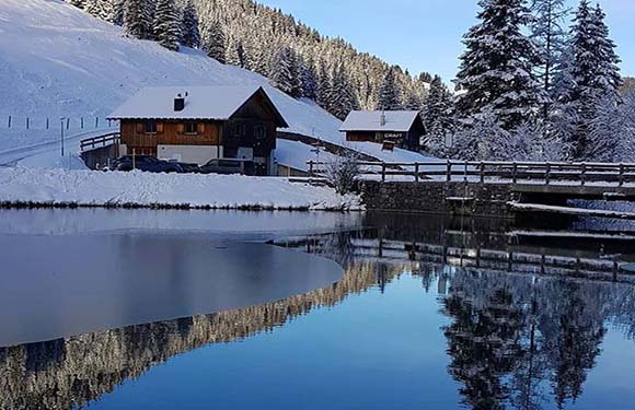 A winter scene with snow-covered ground and trees, featuring a wooden chalet next to a calm lake that reflects the surrounding scenery.