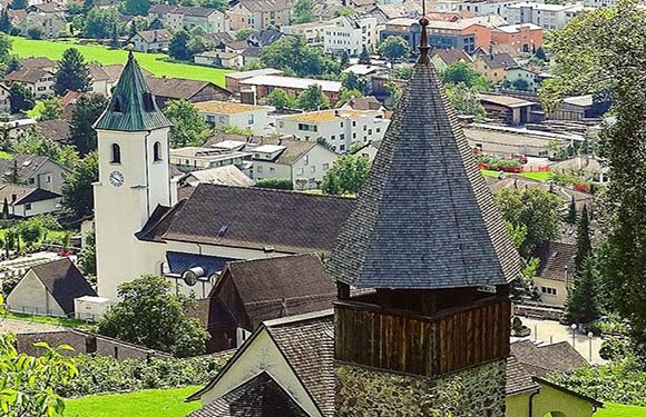 A picturesque view of a European village with traditional buildings and church steeples, surrounded by lush greenery.