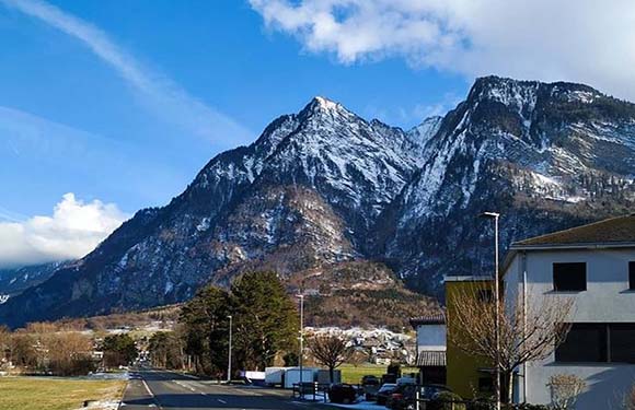 A snowy mountain range under a blue sky, with a building and parking lot in the foreground.