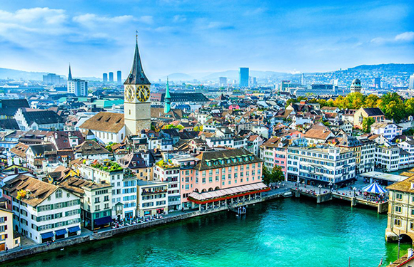 Aerial view of a European city with historical buildings, a prominent church tower, and a river flowing through the center.