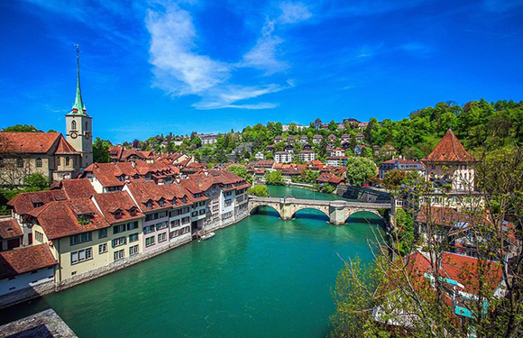 A scenic European town with historic buildings, a church spire, a bridge, and greenery under a blue sky.