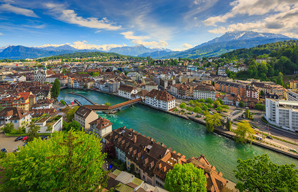 A scenic city with traditional European architecture, crossed by a river and bridges, set against mountains and greenery.