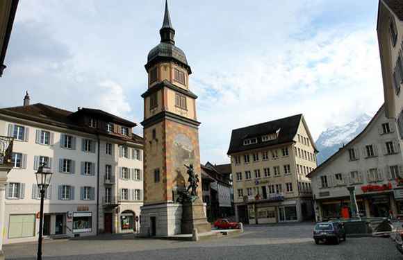 A town square with a clock tower, traditional buildings, and a statue.