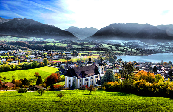 A vivid scene of a white church amid green fields, a town by a lake, and mountains under a partly cloudy sky.