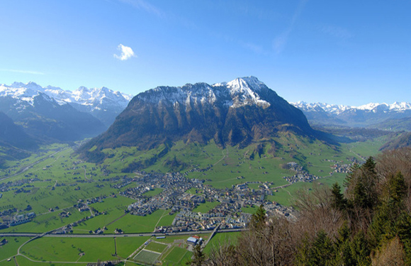 A panoramic view of a lush valley with a town spread out at the base of a prominent mountain, under a clear blue sky with distant snow-capped peaks.