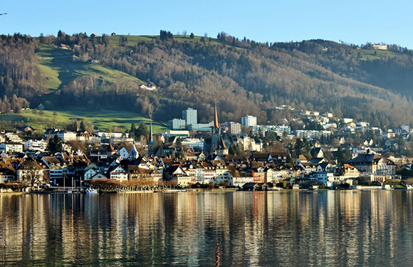 A scenic view of a lakeside town with buildings reflecting on the water, backed by green hills and a clear sky.