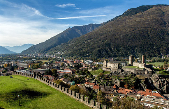 A picturesque town with historic buildings and a fortress, set in a valley with green fields and mountains under a cloudy sky.