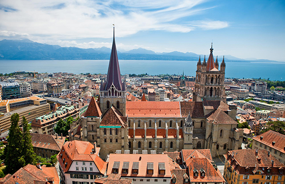 Aerial view of a city featuring historical buildings, a cathedral with two spires, near water and mountains.