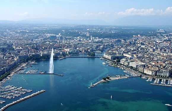 Aerial city view with a lake, water jet fountain, buildings, greenery, and a boat-filled marina on the left.