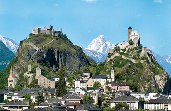 A historic town with old buildings, a hilltop fortress and church, set against snowy mountains.