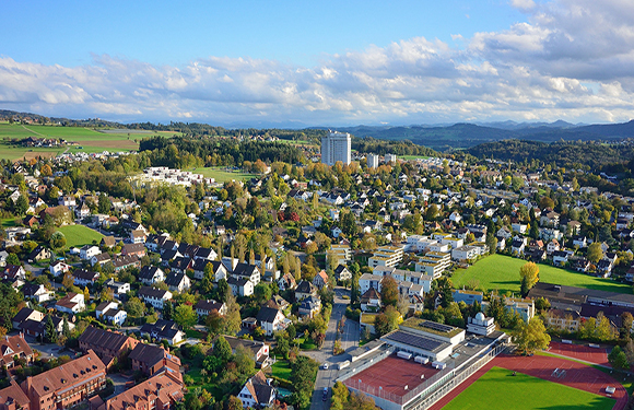 Aerial view of a suburban area with houses, trees, and a sports field under a partly cloudy sky.