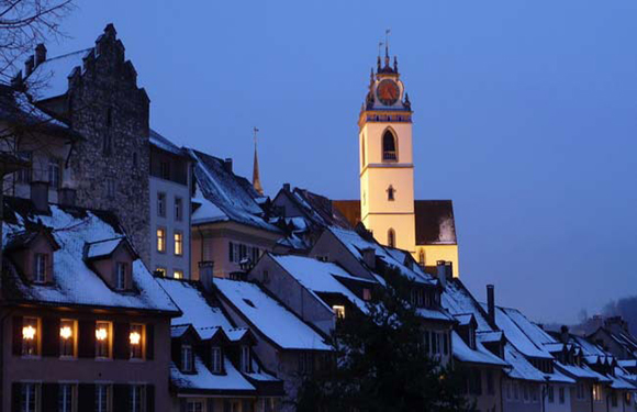 A twilight view of a snow-covered European town with traditional buildings and a prominent church tower illuminated in the background.