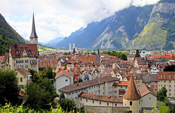 A scenic European town with traditional architecture and a church spire, surrounded by green mountains under a partly cloudy sky.