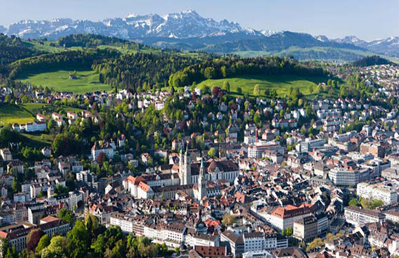 Aerial shot of a European city with dense buildings, a church spire, greenery, and snowy mountains in the background.