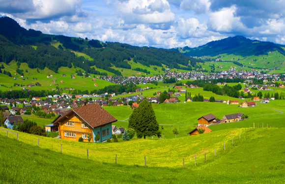 A picturesque landscape featuring a lush green valley with a small town, traditional houses, and rolling hills under a partly cloudy sky.