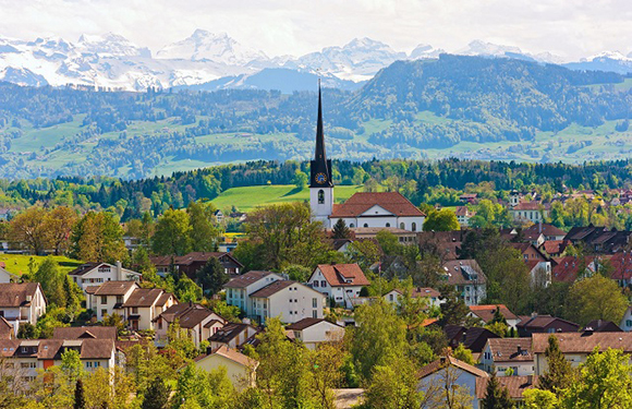 A picturesque European village featuring a church spire, homes, greenery, and snow-capped mountains under a partly cloudy sky.