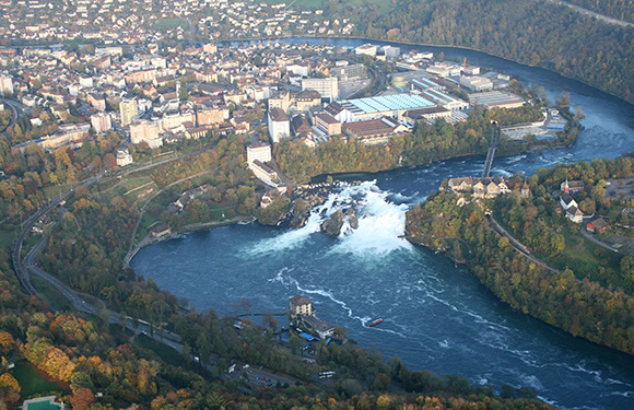 Aerial view of a river with a large waterfall in the center, surrounded by greenery and buildings in the distance.