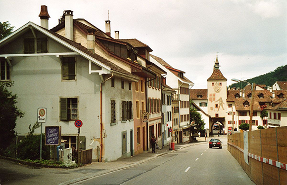 A European town street view with traditional buildings, a distant clock tower, a driving car, and construction fencing on the right.