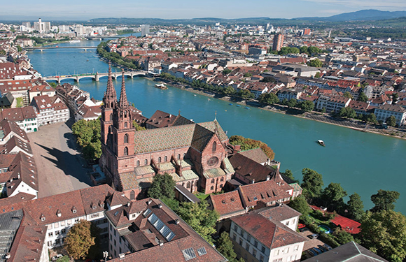 Aerial city view with a river, a red-roofed church, many buildings, and distant hills.