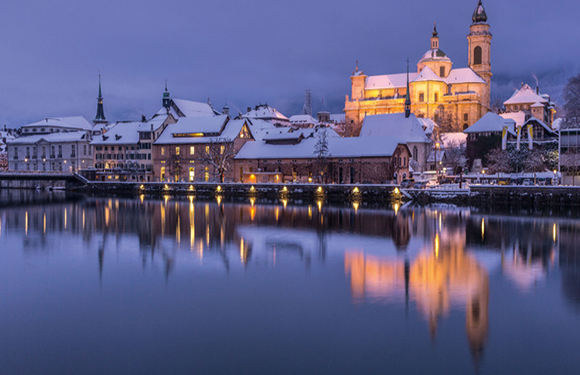 A snowy European town with historic buildings, reflected in calm waters under a warm, glowing dusk sky.