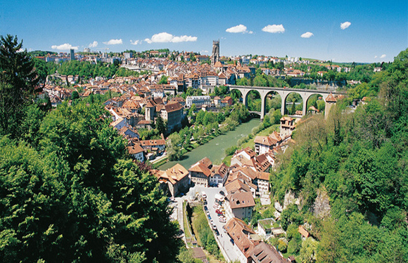 A panoramic view of a historic city featuring traditional buildings, a large arched bridge, a river, and lush greenery under a blue sky.