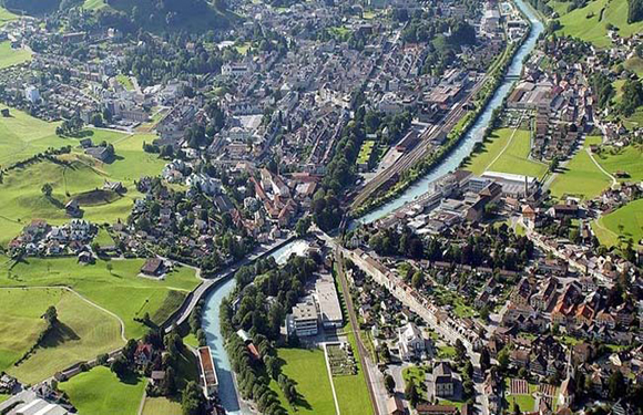 Aerial view of a town with dense buildings alongside a winding river, surrounded by green fields and trees.