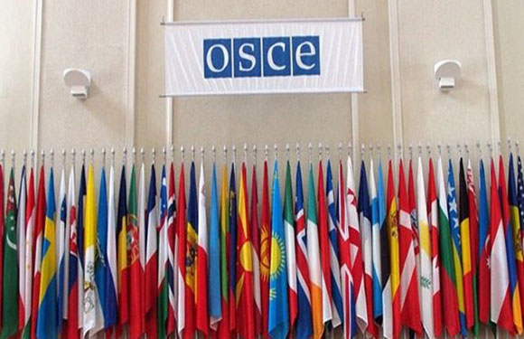 A banner with the letters "OSCE" hangs above a display of various national flags arranged in multiple rows on flagpoles.