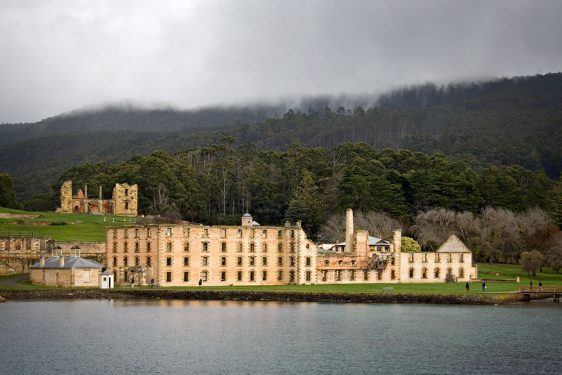 A historic building with multiple chimneys and windows, situated beside a body of water with hills and overcast skies in the background.