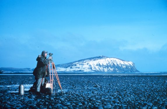 A person using surveying equipment on a rocky terrain with a snow-capped hill in the background under a blue sky.