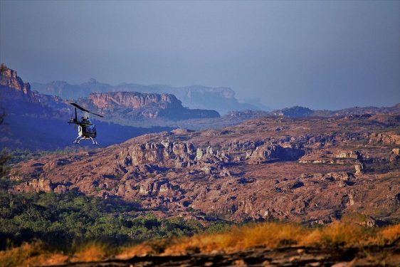 A helicopter flying over a rugged, rocky landscape with hills in the background under a hazy sky.