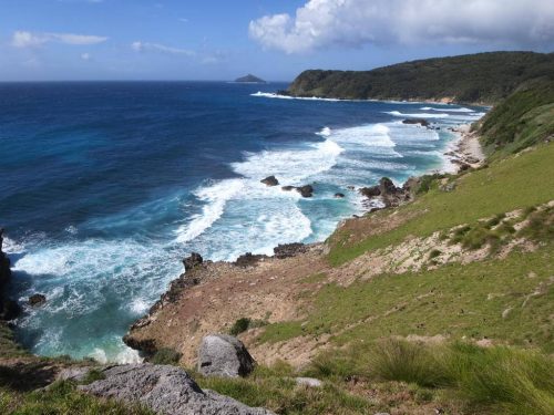 A coastal landscape with waves crashing against rocky shores, a grassy hillside in the foreground, and a clear blue sky with scattered clouds.