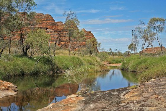 A peaceful landscape with a calm water body, rocky outcrops, greenery, under a blue sky with clouds.