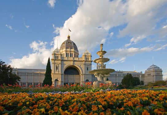 A historical building with a central dome, two smaller domes, wings with arched windows, a garden with orange flowers and a fountain.