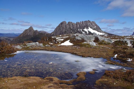 A landscape with a rugged, snowy mountain, a semi-frozen pond, and a blue sky with scattered clouds.