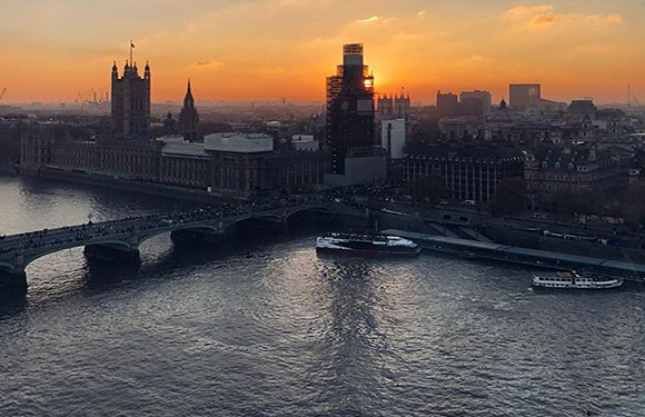 A sunset view over a city with a river, showing a bridge, buildings, and a boat on the water.