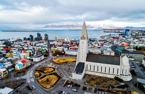 Aerial view of a cityscape with colorful buildings, a prominent church with a tall spire, and a coastline with mountains in the distance.