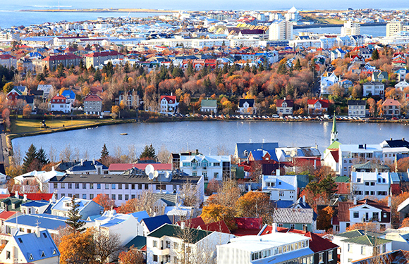 Aerial view of a colorful cityscape with dense buildings near a body of water, trees in autumn hues, and a clear sky.