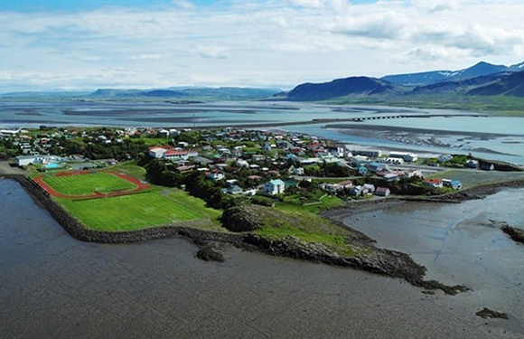 Aerial view of a coastal town with buildings and streets, surrounded by water and green landscapes, under a partly cloudy sky.