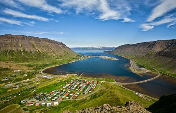 A picturesque small town in a valley with vibrant buildings, encircled by mountains, overlooking a serene blue fjord.