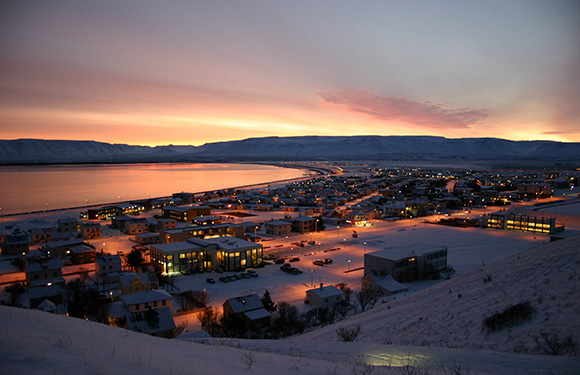 A scenic view of a coastal town at dusk with lights from buildings reflecting on the water, under a sky with hues of orange and pink.