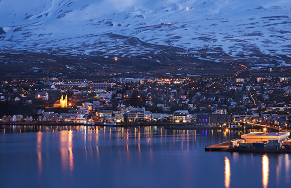 A nighttime cityscape with illuminated buildings reflected in a body of water, with snow-covered mountains in the background.