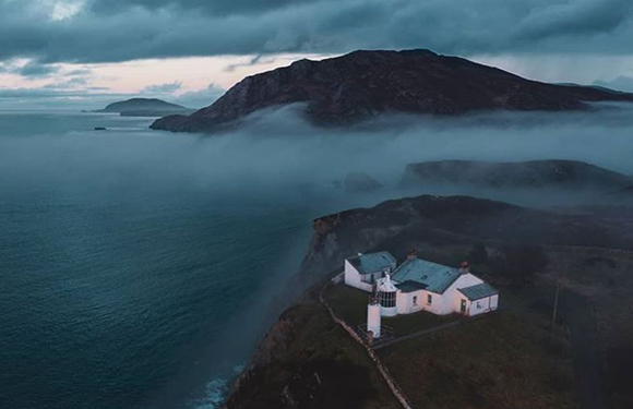 Aerial view of a coastal landscape at dusk with low-lying clouds, featuring a small cluster of buildings on a promontory overlooking the sea.