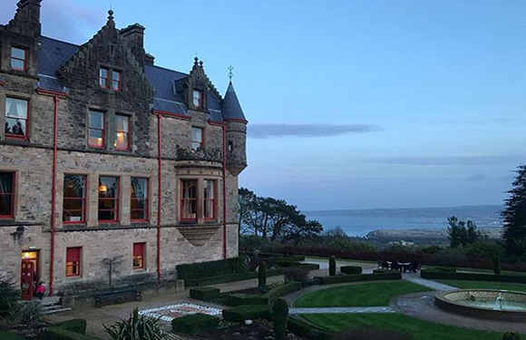 An image of a grand historic building at dusk with lit windows, manicured gardens in the foreground, and a view of the sea in the background.