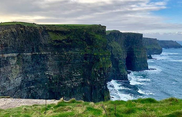 A scenic view of the Cliffs of Moher with lush green grass in the foreground and the Atlantic Ocean below under a cloudy sky.